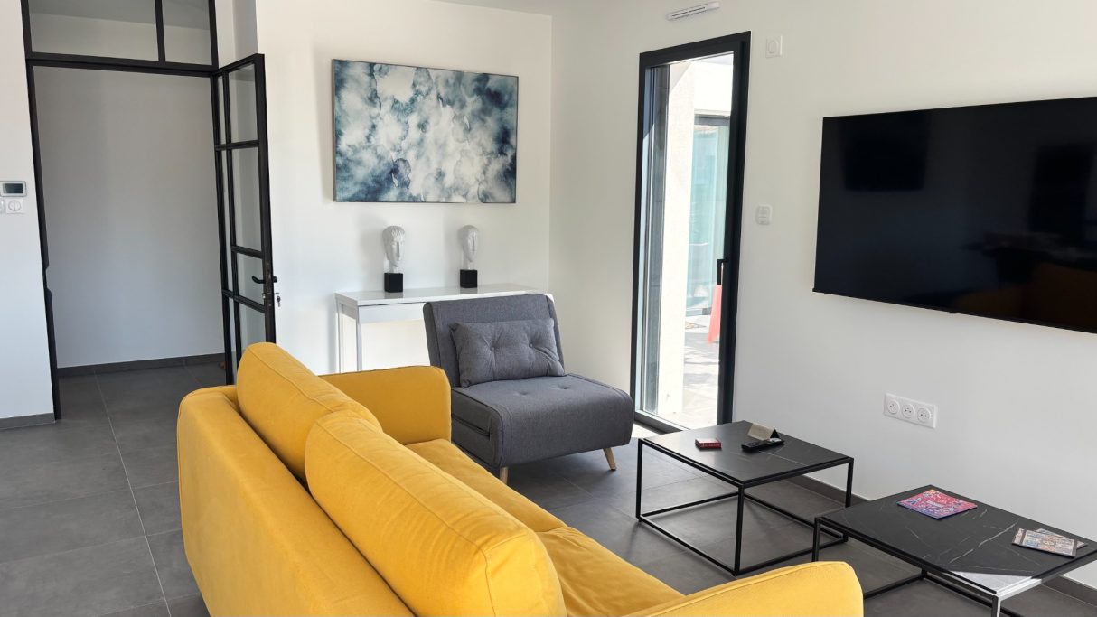 Salon moderne avec un canapé jaune, fauteuil gris, tables noires et TV murale.