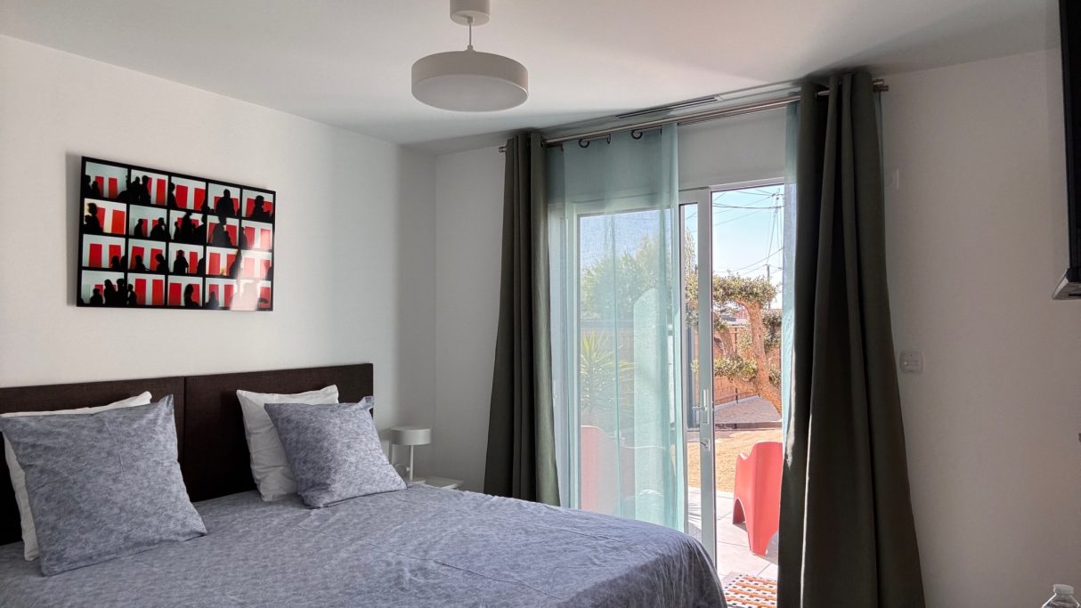 Chambre lumineuse avec lit double, rideaux gris et fenêtre donnant sur l’extérieur.